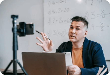 An individual seated at a desk in front of an open laptop, gesturing towards a whiteboard filled with algebraic equations while being recorded by a camera on a tripod.